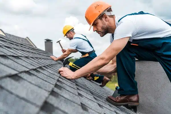 Two roofers in hard hats installing shingles on a house.